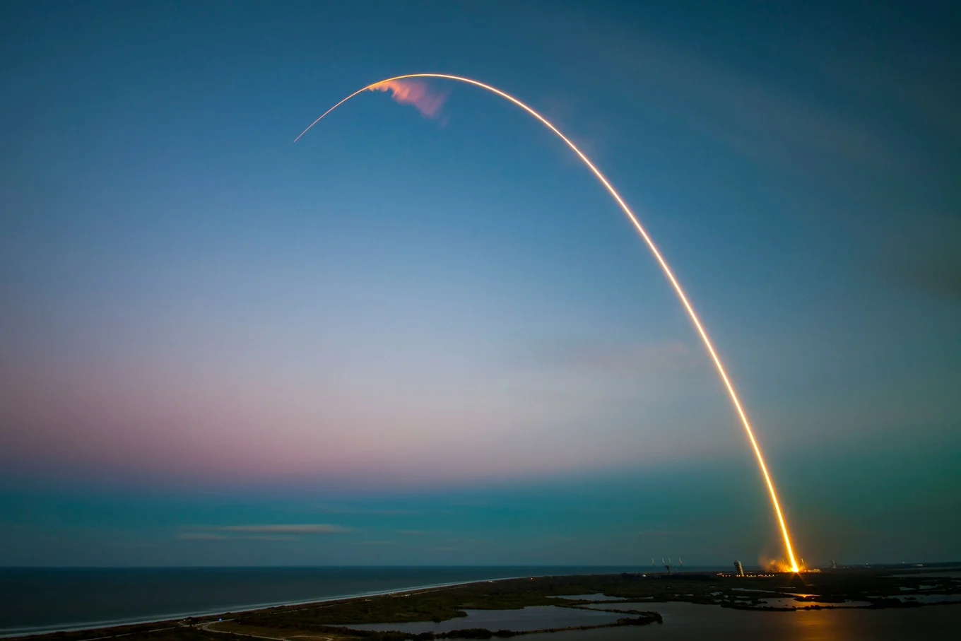 A zoomed-out shot of a shoreline in twilight, with an arc of light stretching from the lower right corner to the top left, it's the trail of a rocket launch.
